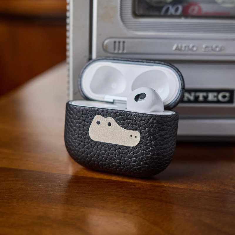 Premium leather AirPods Pro cover in emoji design Black leather case with a logo containing white earbuds on a wooden surface with a vintage radio in the background.
