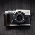 A silver and black Om System Pen camera with a dark wood grip attachment and a black lens, photographed from above on a dark textured background.
